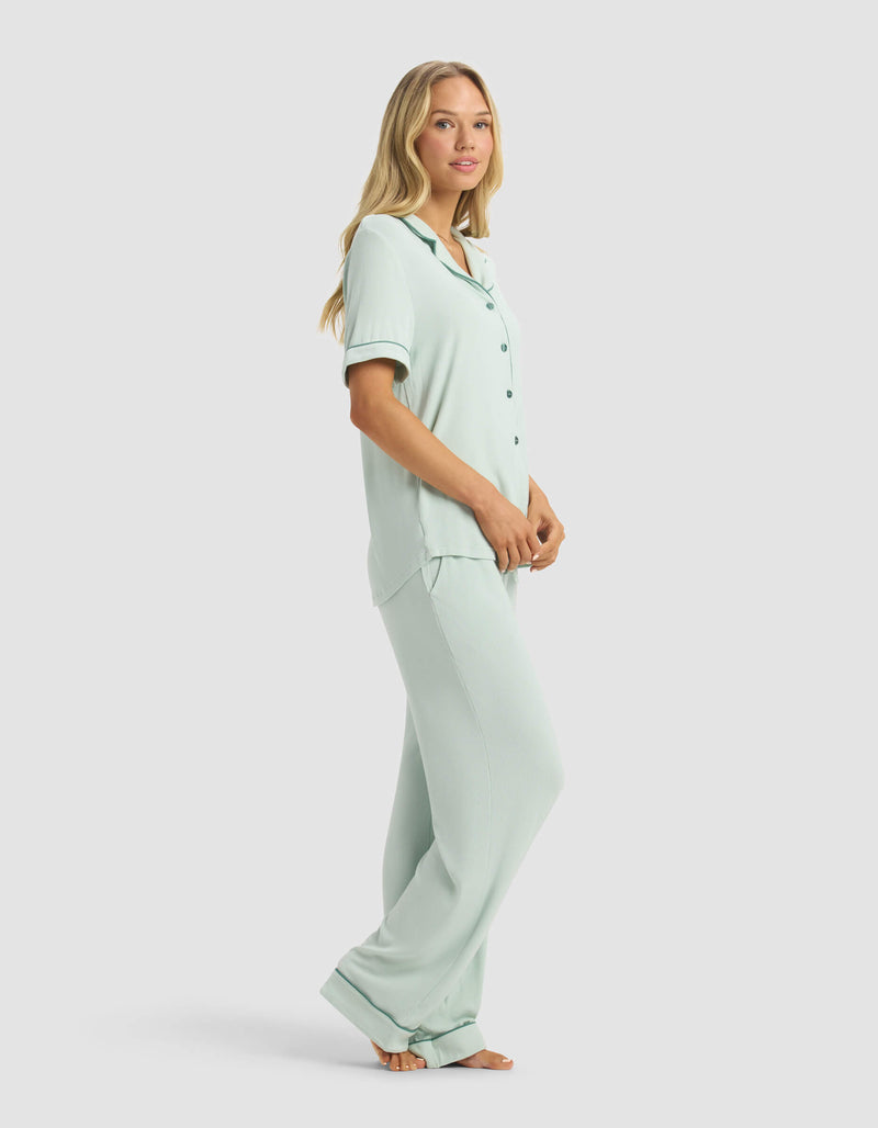 A woman stands barefoot in the Cozy Earth Women's Rib-Knit Classic Short Sleeve & Pant Pajama Set, featuring a light blue button-up top and pants. She poses side-on, looking at the camera against a plain light background.