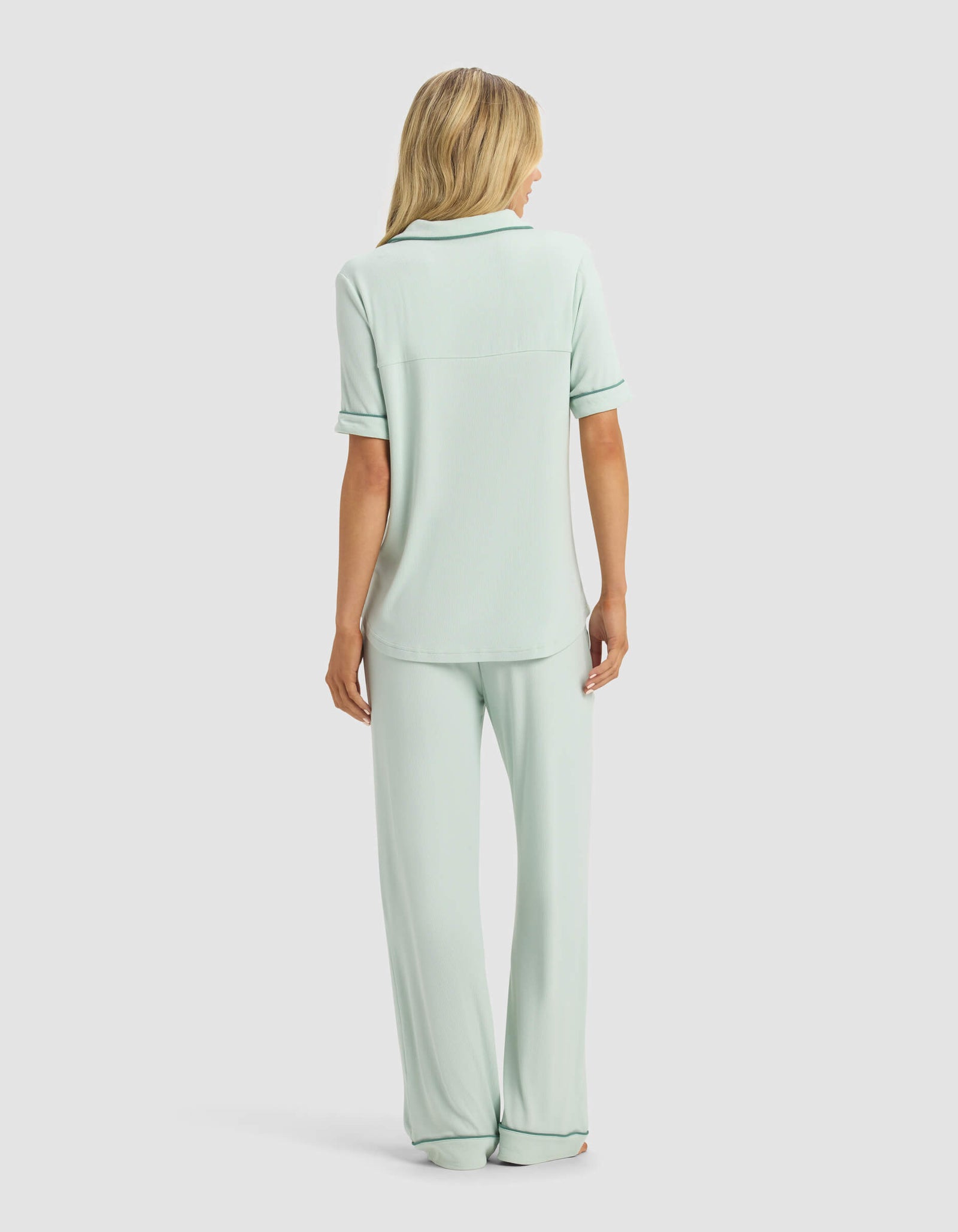 A blonde woman faces away in a Cozy Earth Women's Rib-Knit Classic Short Sleeve & Pant Pajama Set, featuring a light green color, short sleeves, collar, and long pants. The background is plain and light-colored.