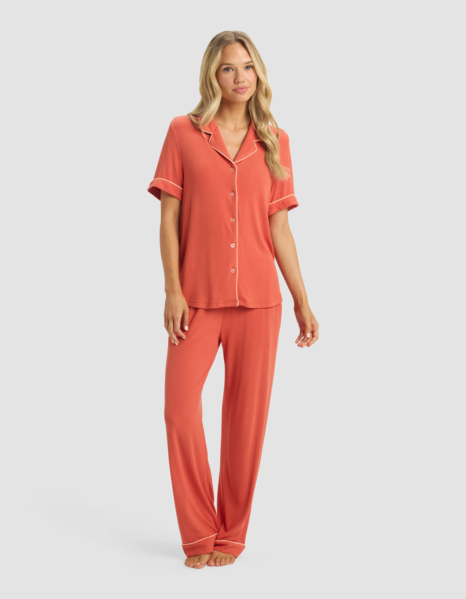 A woman models the Cozy Earth Women's Rib-Knit Classic Short Sleeve & Pant Pajama Set in coral, featuring a short-sleeve button-up top and long pants, standing barefoot against a light gray background.