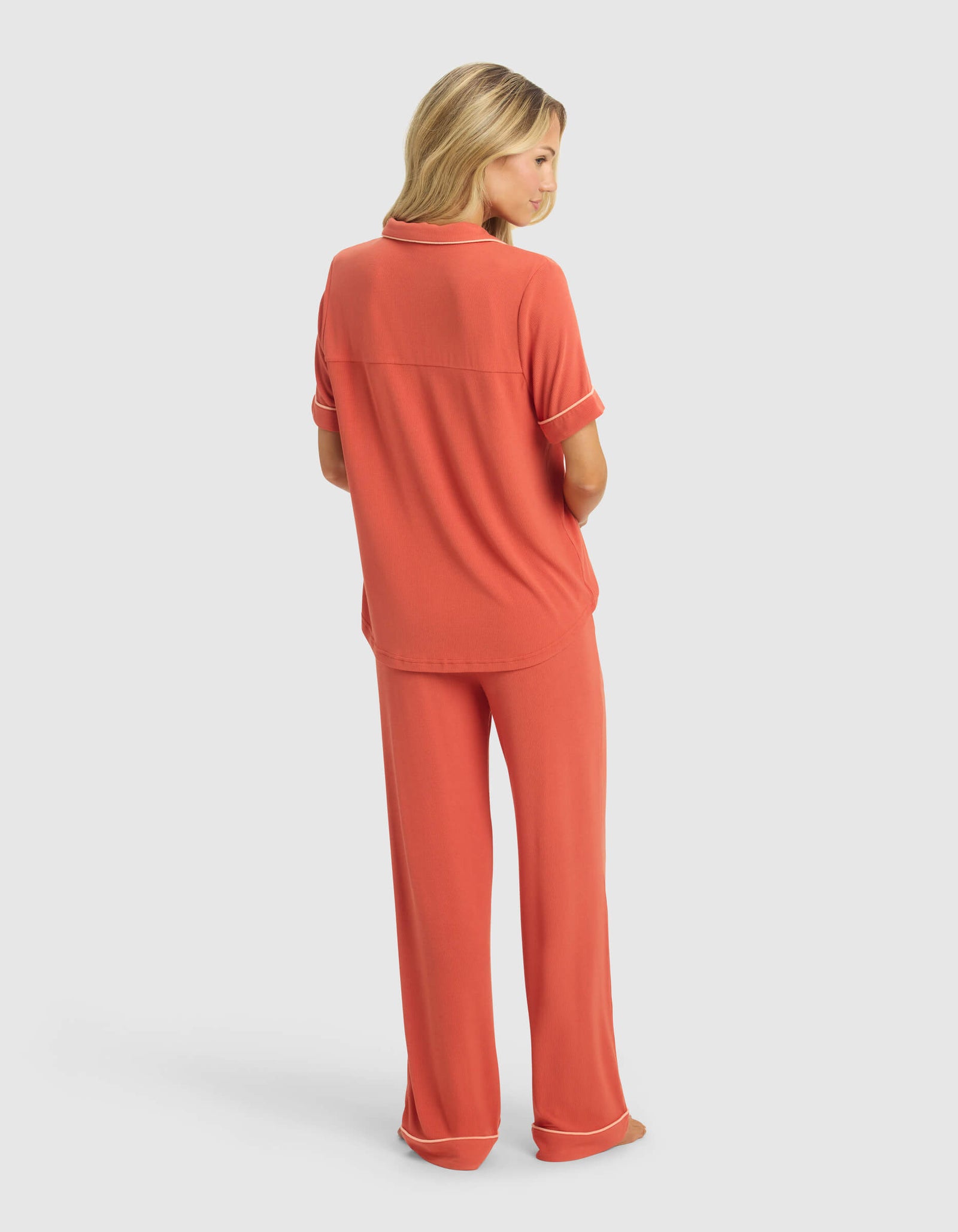 A woman with long blonde hair faces away, wearing Cozy Earth's Women's Rib-Knit Classic Short Sleeve & Pant Pajama Set in coral with cream piping at the cuffs and hem, standing against a plain white background.