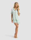 A woman stands barefoot against a light background, wearing the Cozy Earth Women's Bamboo Rib-Knit Classic Short Sleeve Pajama Set in light green, smiling gently as she touches her hair with one hand.|Color:Seafoam