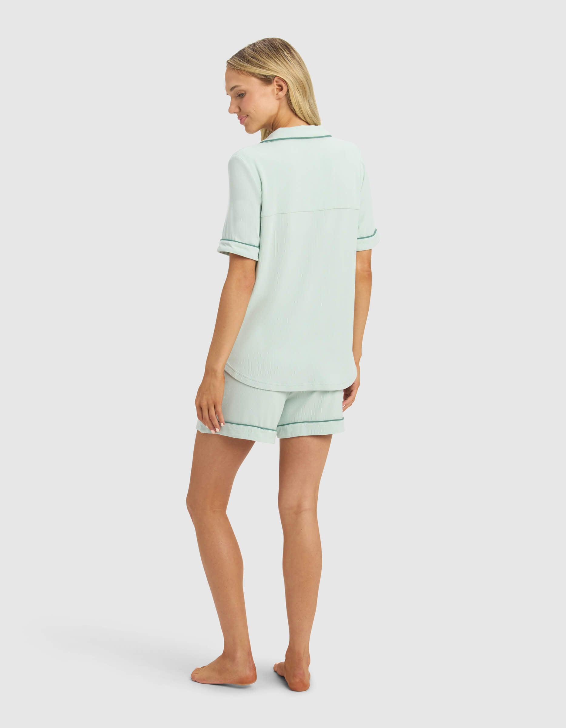 A woman stands barefoot, facing away, wearing the Cozy Earth Women's Bamboo Rib-Knit Classic Short Sleeve Pajama Set with matching shorts and dark trim, in front of a plain light background.|Color:Seafoam