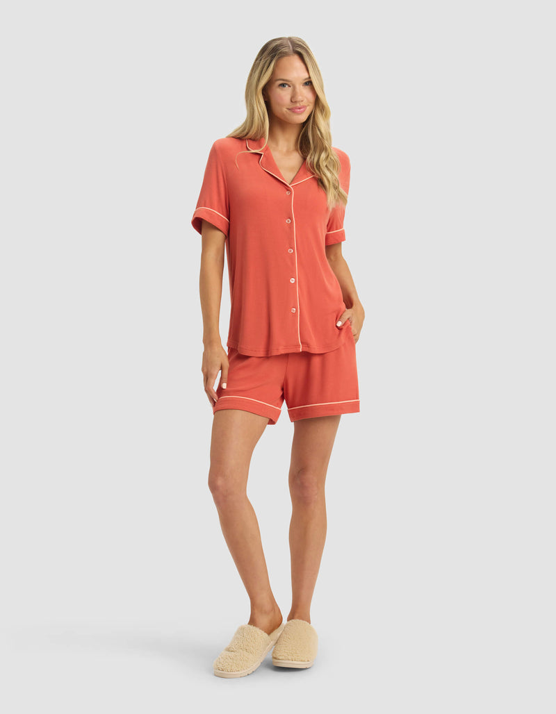 A woman with long blonde hair models the Cozy Earth Women's Bamboo Rib-Knit Classic Short Sleeve Pajama Set in coral with white trim and matching shorts, paired with beige fuzzy slippers, against a plain light background.