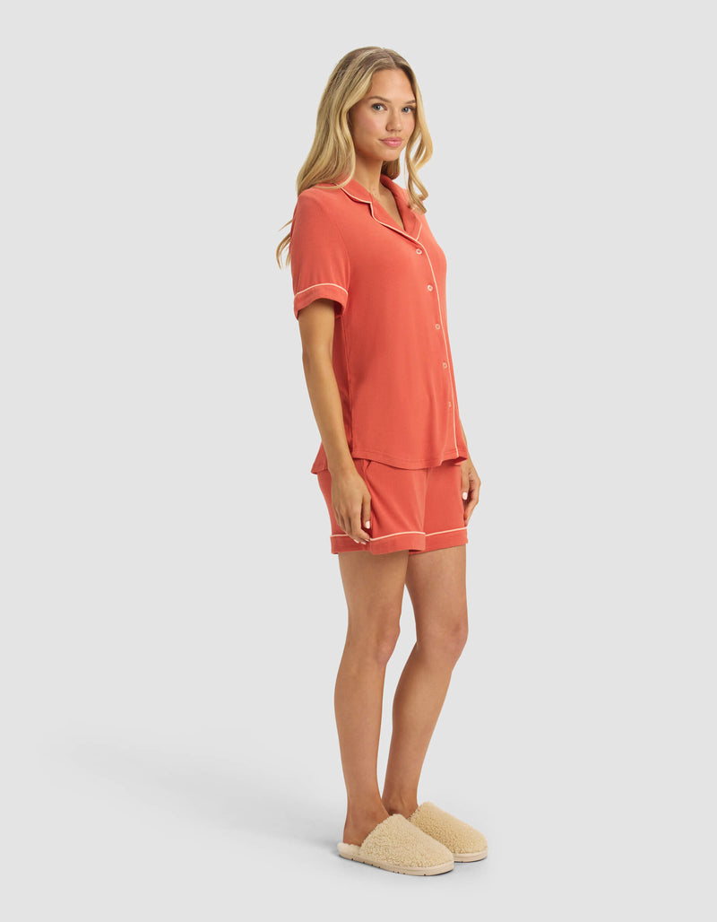 A woman wears the Cozy Earth Women's Bamboo Rib-Knit Classic Short Sleeve Pajama Set in coral with white trim, paired with beige fuzzy slippers, standing against a plain light gray background.