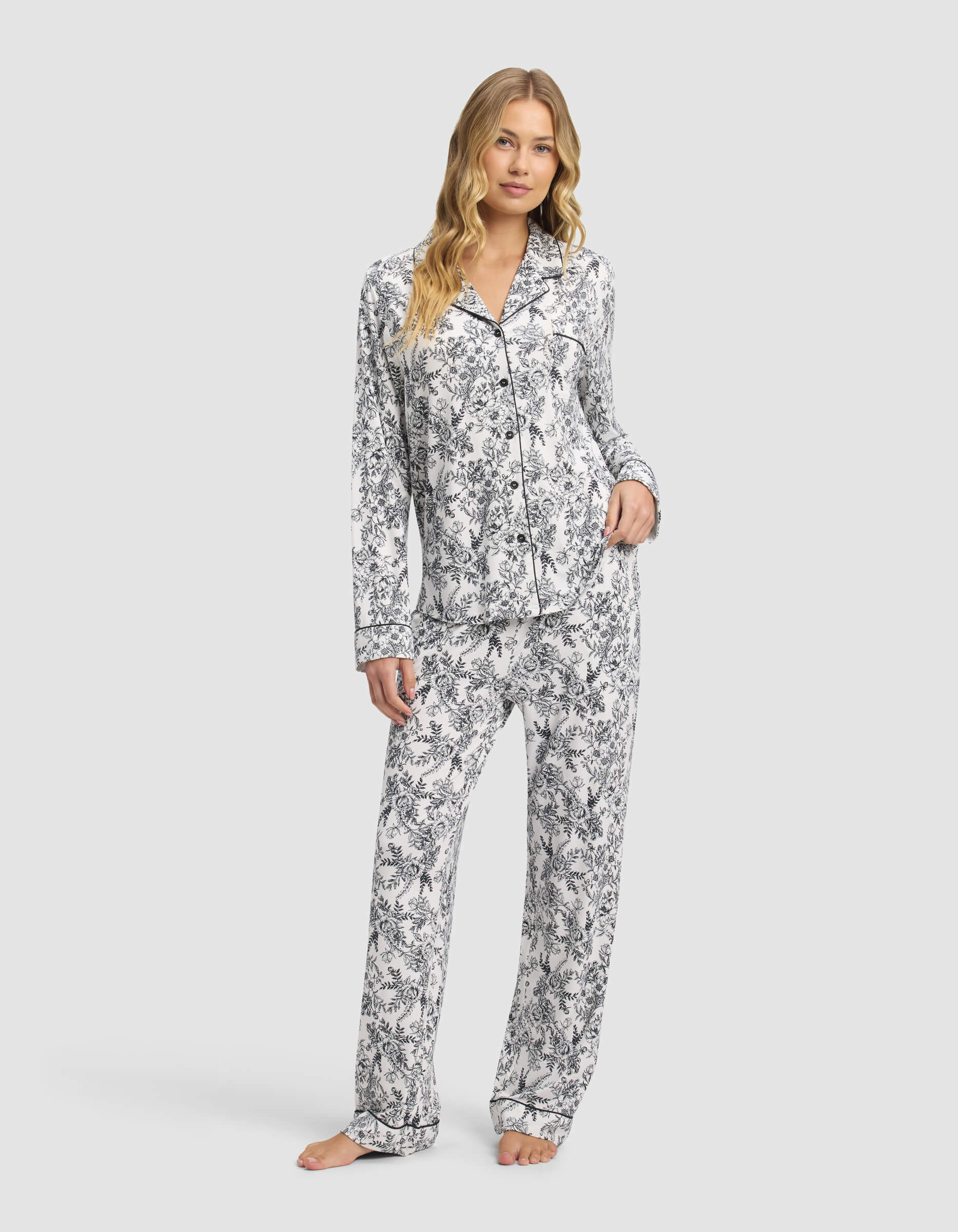 A woman with long wavy hair and a relaxed expression stands barefoot, wearing Cozy Earth's Women's Bamboo Stretch-Knit Long Sleeve Pajama Set featuring a black and white floral pattern with a button-up top and loose pants.|Color:Jet Black Toile