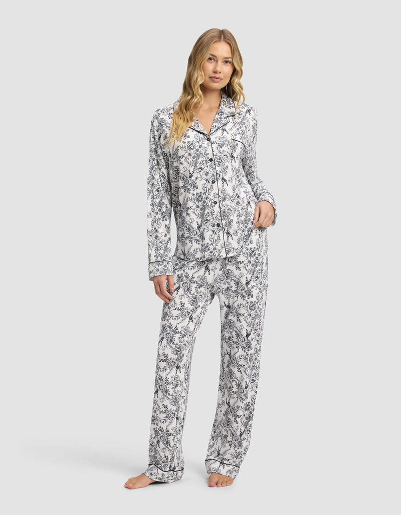 A woman with long wavy hair and a relaxed expression stands barefoot, wearing Cozy Earth's Women's Bamboo Stretch-Knit Long Sleeve Pajama Set featuring a black and white floral pattern with a button-up top and loose pants.