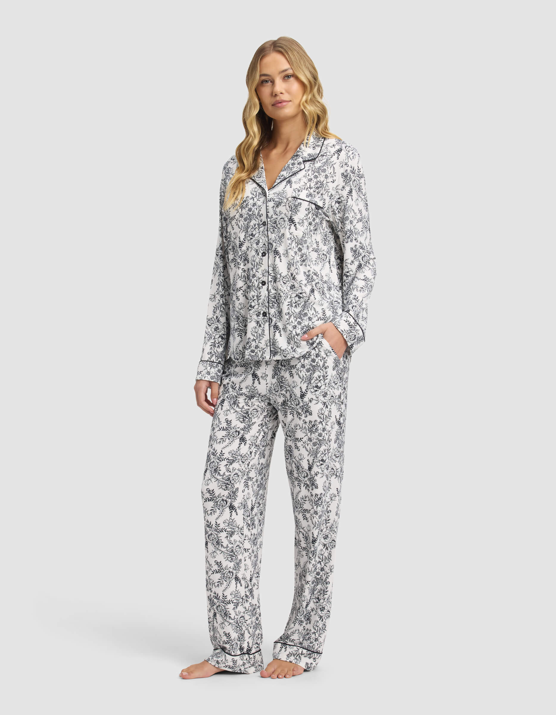 A woman with long, wavy blonde hair wears the Cozy Earth Women's Bamboo Stretch-Knit Long Sleeve Pajama Set in white with a black floral pattern. The collared, button-up top and loose pants create a relaxed look as she stands barefoot against a light background.|Color:Jet Black Toile