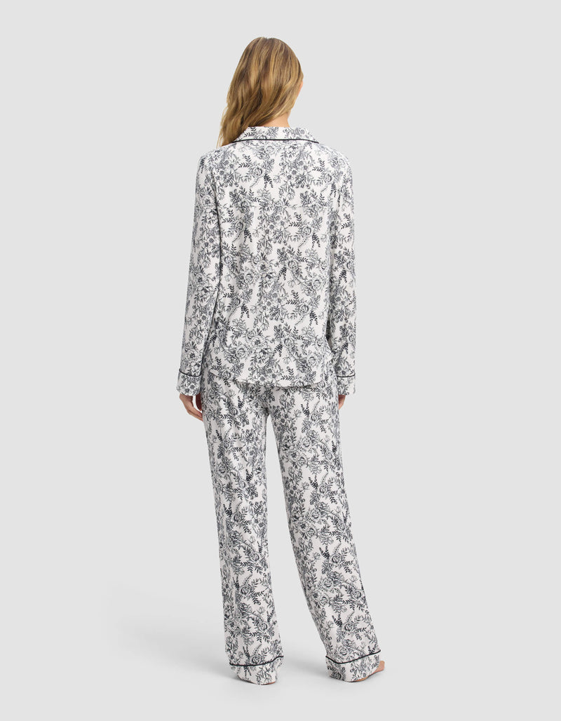 A woman with long, wavy blonde hair faces away, wearing the Cozy Earth Women's Bamboo Stretch-Knit Long Sleeve Pajama Set in white with a black floral pattern, against a plain light gray background.