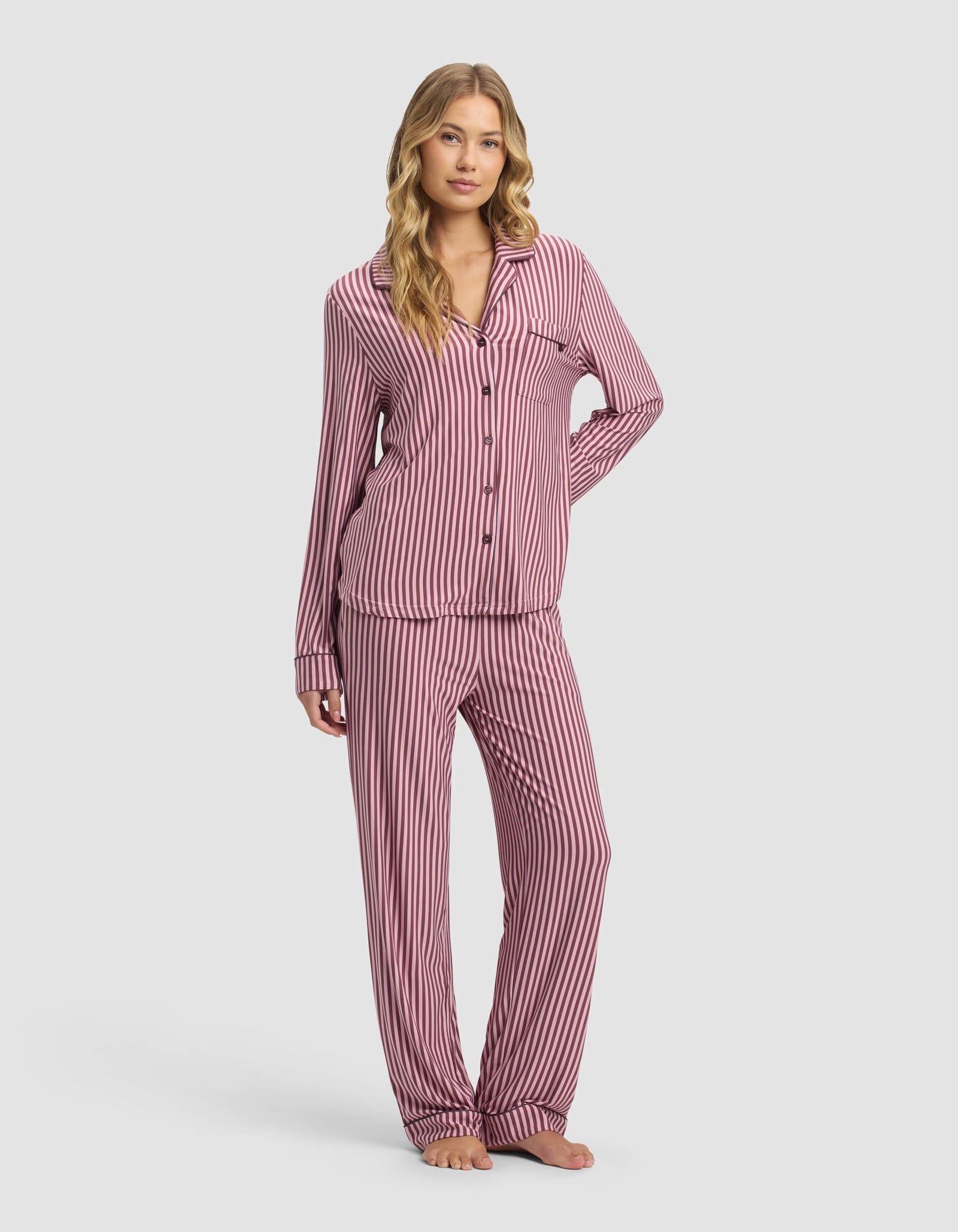 A woman stands barefoot against a plain background, wearing the Cozy Earth Women's Bamboo Stretch-Knit Long Sleeve Pajama Set—featuring pink and white vertical stripes with a button-up top and loose pants.