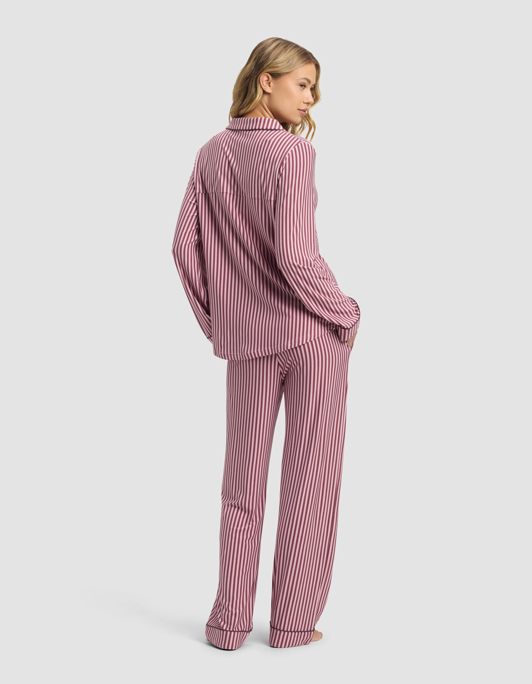 A woman stands barefoot, facing away, wearing the Cozy Earth Women's Bamboo Stretch-Knit Long Sleeve Pajama Set in pink and white stripes. The background is plain and light-colored.|Color:Market Stripe in Mulberry