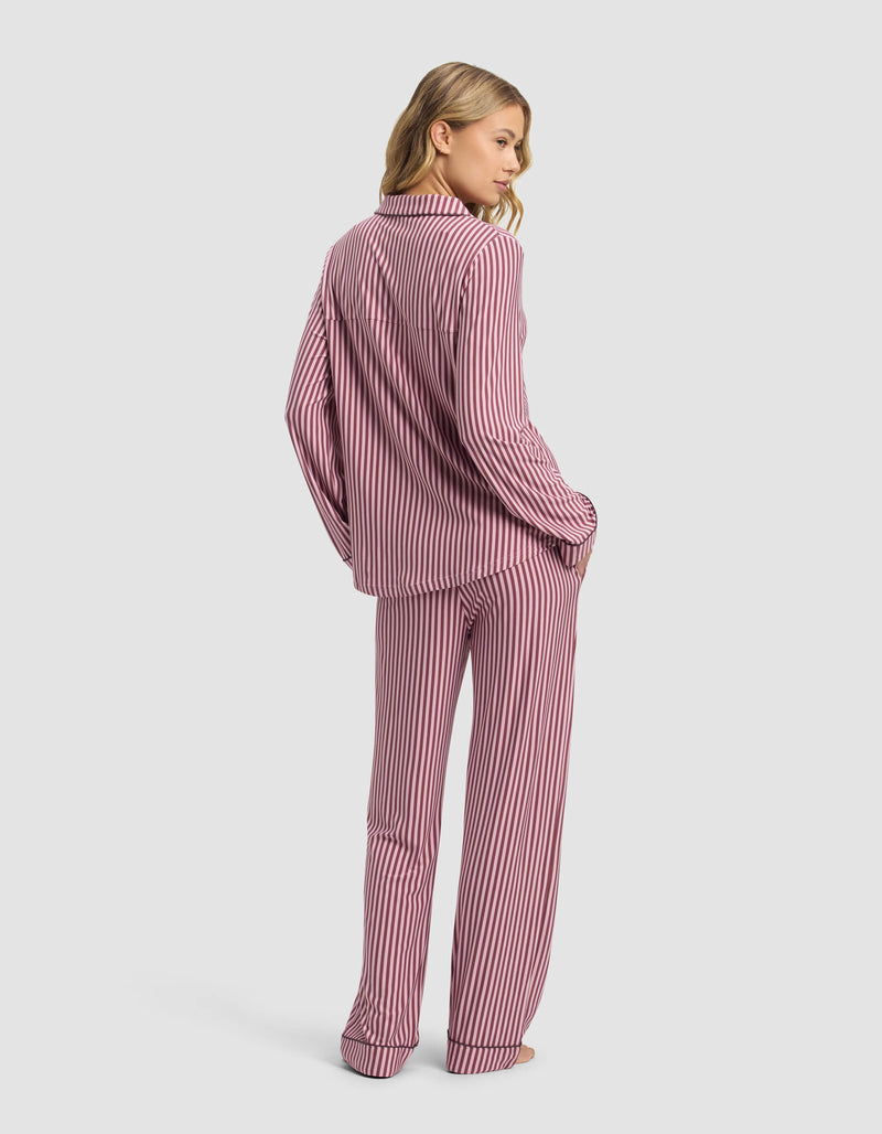 A woman stands barefoot, facing away, wearing the Cozy Earth Women's Bamboo Stretch-Knit Long Sleeve Pajama Set in pink and white stripes. The background is plain and light-colored.
