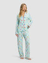 A woman with long blonde hair models the Cozy Earth Women's Bamboo Stretch-Knit Long Sleeve Pajama Set—featuring a button-up top and loose pants in light blue with a colorful heart pattern—standing barefoot against a plain background.|Color:Seafoam Sleepy Hearts