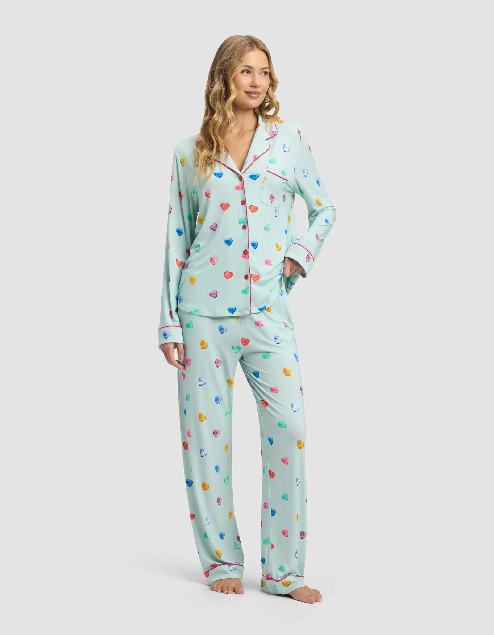 A woman with long blonde hair models the Cozy Earth Women's Bamboo Stretch-Knit Long Sleeve Pajama Set—featuring a button-up top and loose pants in light blue with a colorful heart pattern—standing barefoot against a plain background.