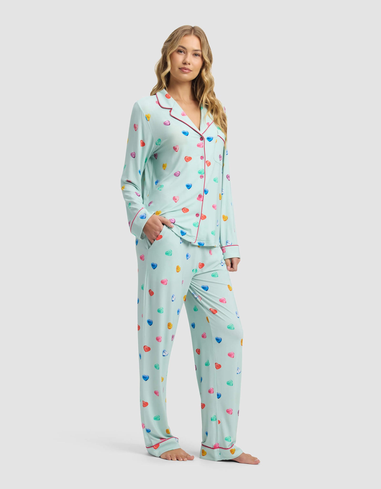 A woman stands barefoot, smiling gently in Cozy Earth's Women's Bamboo Stretch-Knit Long Sleeve Pajama Set—light blue with colorful hearts, red piping, and a button-up shirt. Her hand is in her pocket against a plain light background.