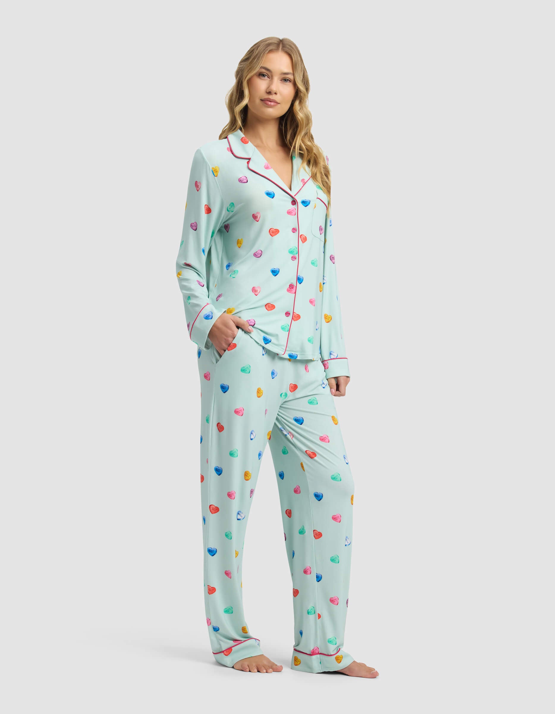 A woman stands barefoot, smiling gently in Cozy Earth's Women's Bamboo Stretch-Knit Long Sleeve Pajama Set—light blue with colorful hearts, red piping, and a button-up shirt. Her hand is in her pocket against a plain light background.|Color:Seafoam Sleepy Hearts