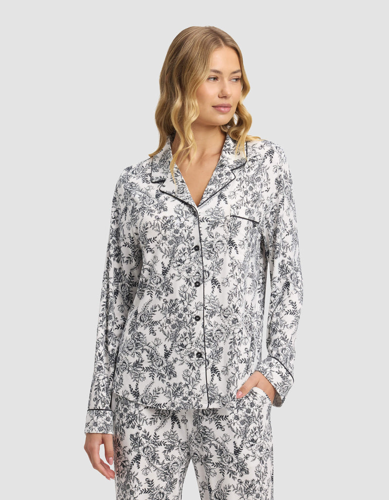 A woman with wavy blonde hair models the Cozy Earth Women's Bamboo Stretch-Knit Long Sleeve Pajama Set, featuring a black and white floral pattern, collared button-up top with chest pocket, as she stands with one hand in her pants pocket.