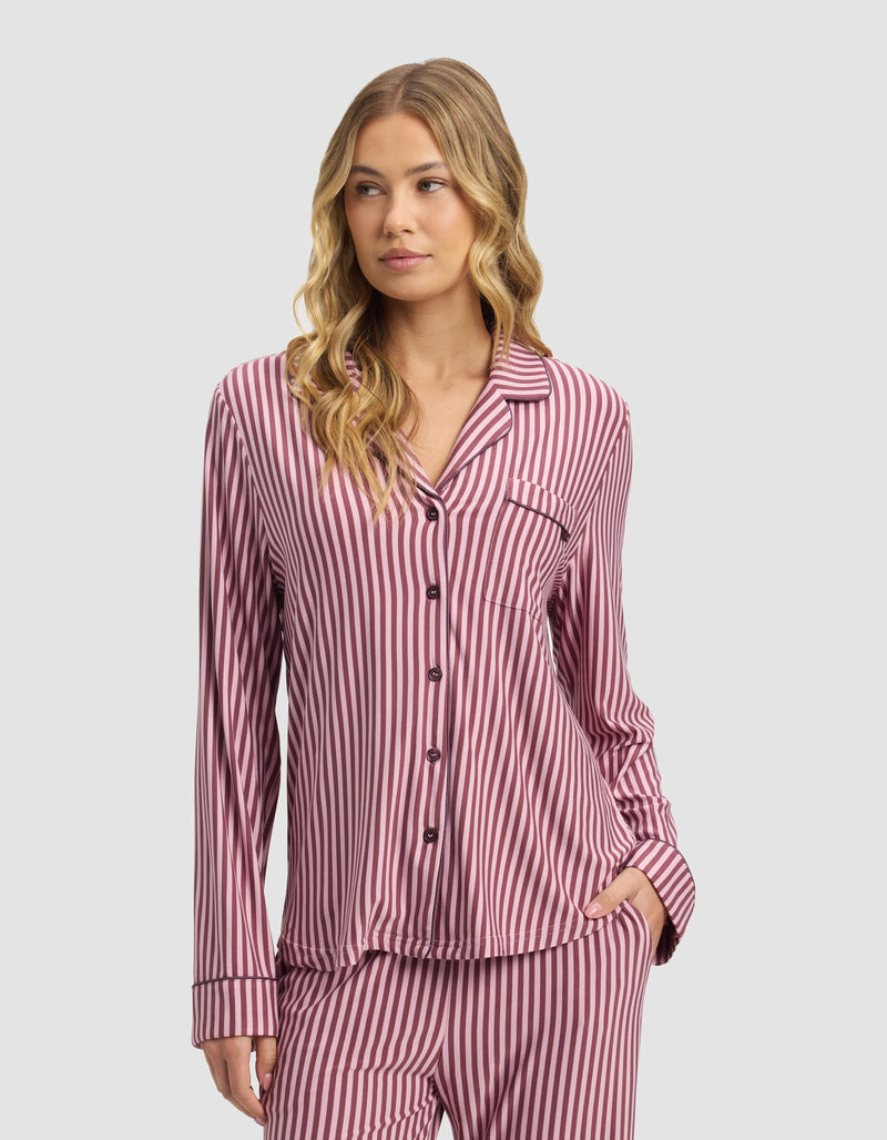 A woman with long wavy blonde hair wears the Cozy Earth Women's Bamboo Stretch-Knit Long Sleeve Pajama Top in pink and white stripes, paired with matching pants, standing against a plain light gray background.