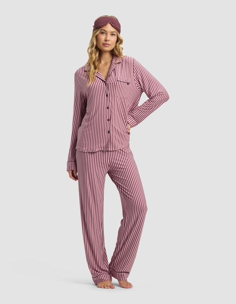 A woman wears the Cozy Earth Women's Bamboo Stretch-Knit Long Sleeve Pajama Top in mauve and white stripes with a matching sleep mask, standing barefoot with one hand on her hip against a plain light background.