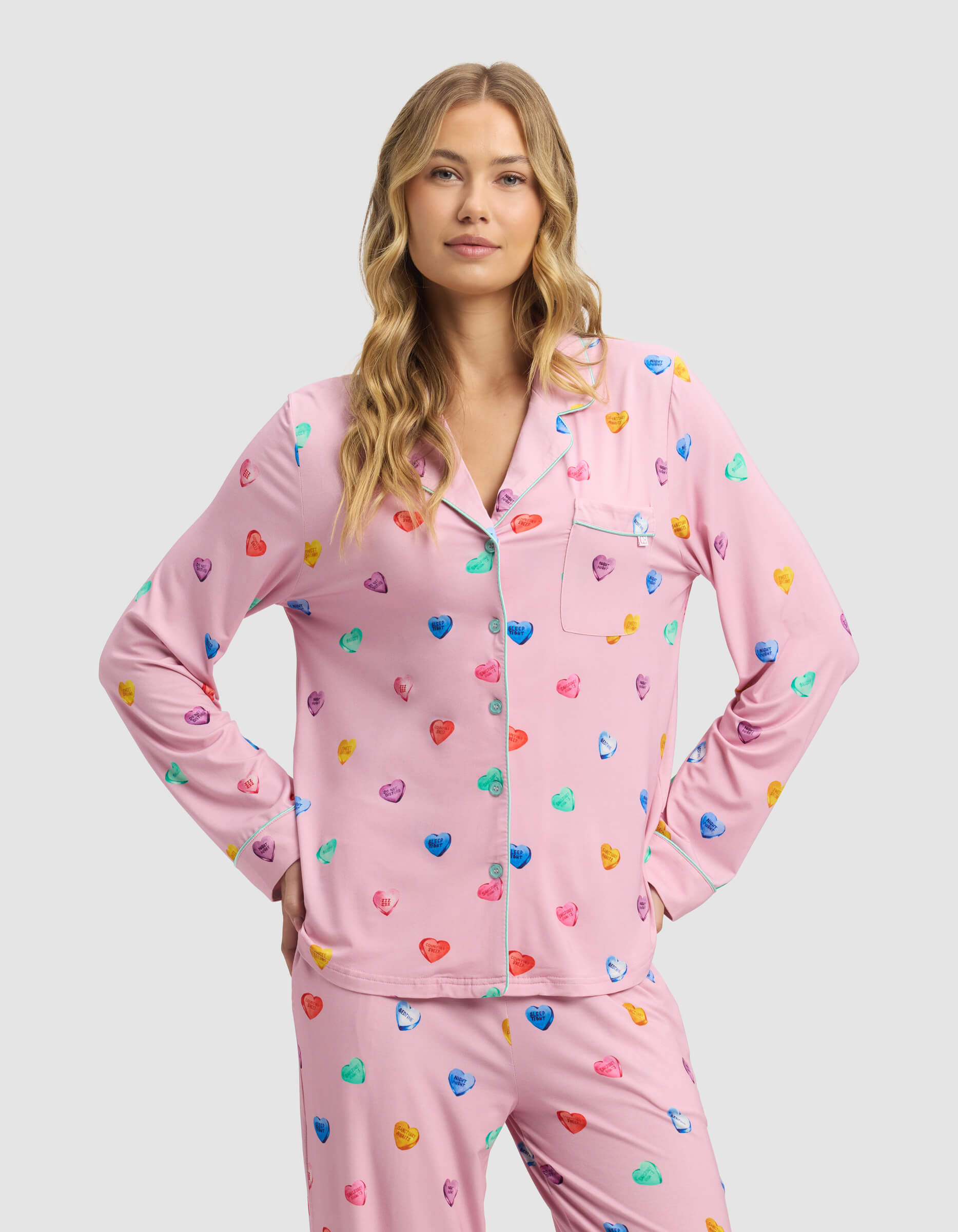A woman with wavy blonde hair wears a pink Women's Bamboo Stretch-Knit Long Sleeve Pajama Top by Cozy Earth, decorated with colorful hearts. She stands with her hands on her hips, smiling slightly against a plain light gray background.