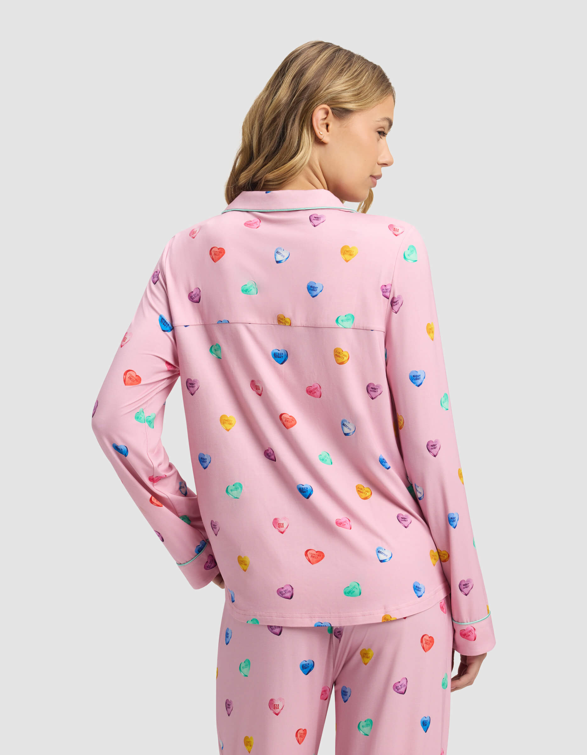 A woman with blonde hair models the Cozy Earth Women's Bamboo Stretch-Knit Long Sleeve Pajama Top in pink with colorful heart patterns, shown from the back and looking over her shoulder against a light gray background.|Color:Primrose Sleepy Hearts