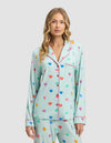 A woman with wavy blonde hair wears the Women's Bamboo Stretch-Knit Long Sleeve Pajama Top by Cozy Earth in light blue with colorful heart patterns and pink trim, standing against a plain light gray background.|Color:Seafoam Sleepy Hearts