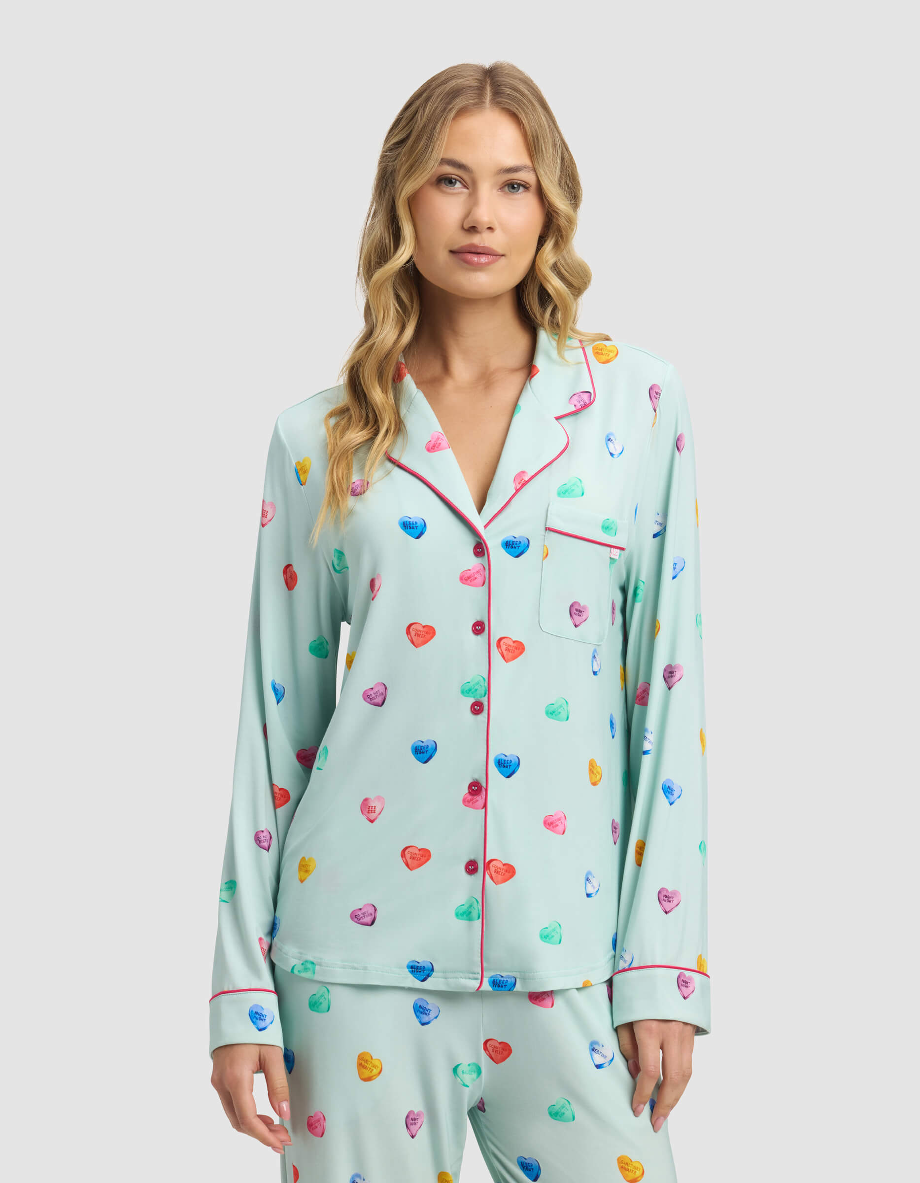 A woman with wavy blonde hair wears the Women's Bamboo Stretch-Knit Long Sleeve Pajama Top by Cozy Earth in light blue with colorful heart patterns and pink trim, standing against a plain light gray background.|Color:Seafoam Sleepy Hearts