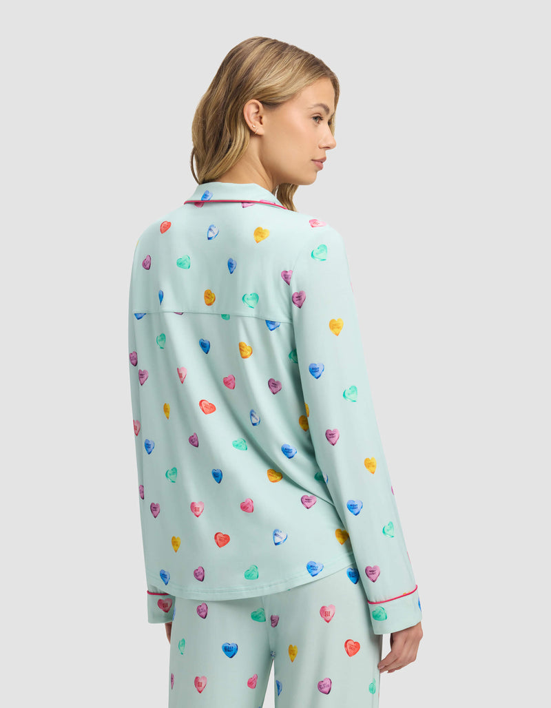 A woman with blonde hair models the back of Cozy Earth's Women's Bamboo Stretch-Knit Long Sleeve Pajama Top, featuring a light blue color and colorful heart patterns. She faces away, turning her head slightly to the side.