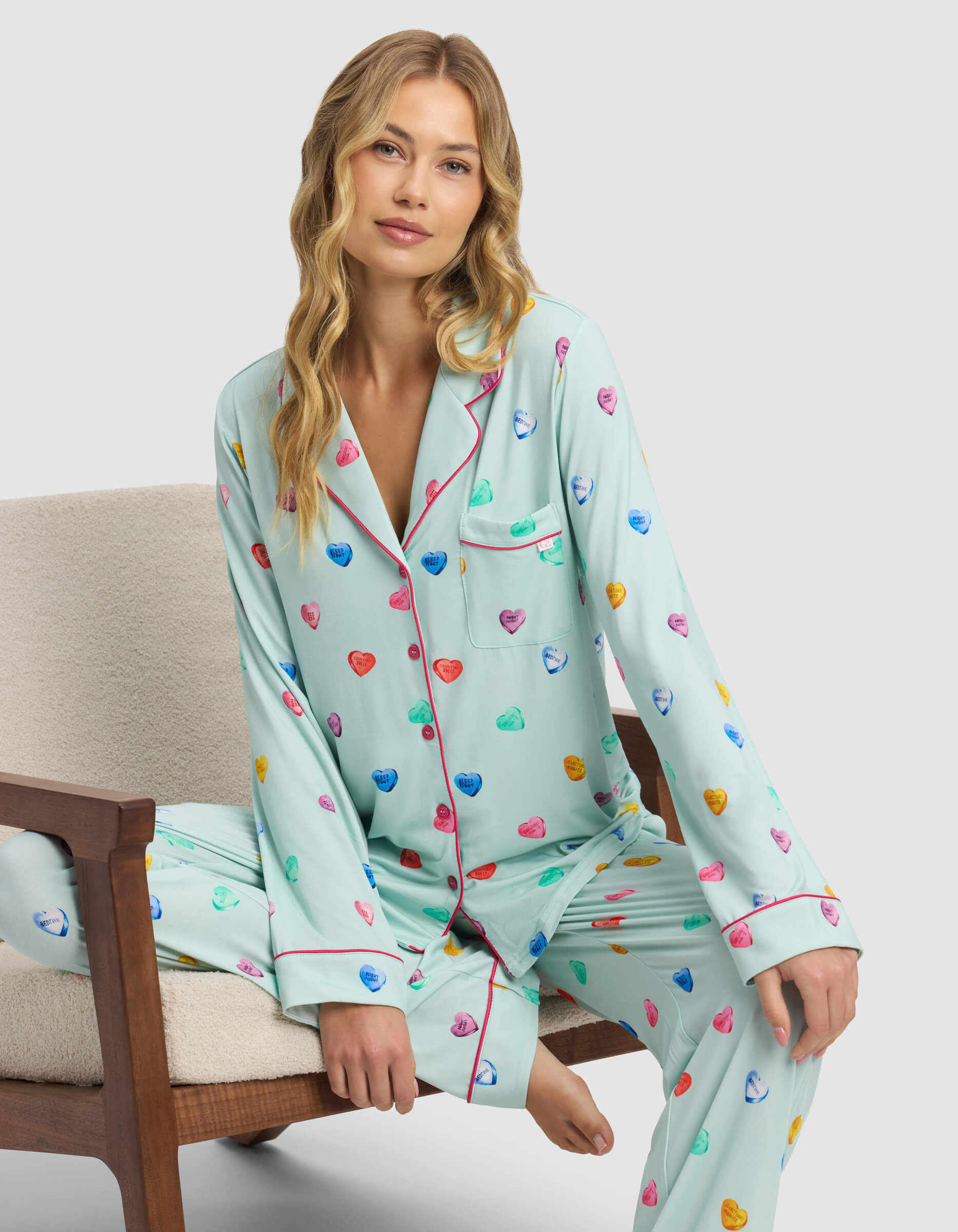 A woman with long blonde hair sits on a beige chair wearing the Cozy Earth Women's Bamboo Stretch-Knit Long Sleeve Pajama Top in light blue, featuring colorful heart patterns and pink piping. She gazes at the camera with a relaxed expression.