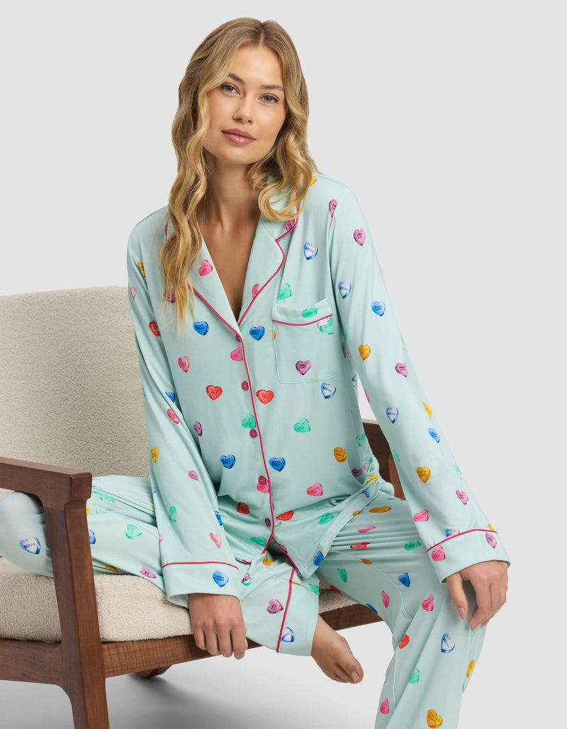 A woman with long blonde hair sits on a beige chair wearing the Cozy Earth Women's Bamboo Stretch-Knit Long Sleeve Pajama Top in light blue, featuring colorful heart patterns and pink piping. She gazes at the camera with a relaxed expression.
