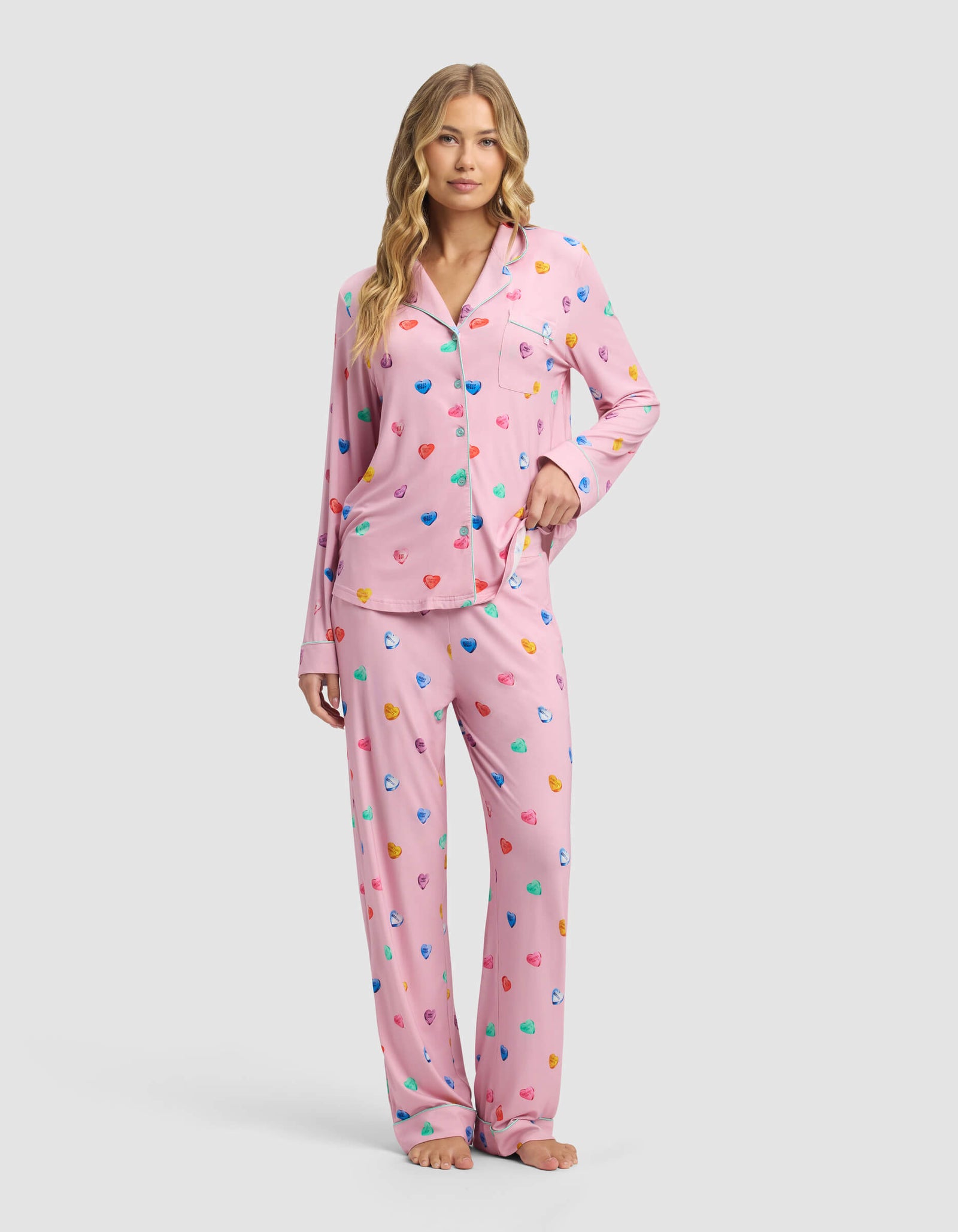 A woman with long, wavy blonde hair and a neutral expression stands barefoot in Cozy Earth’s Women’s Bamboo Stretch-Knit Long Sleeve Pajama Set, featuring pink fabric with a colorful scattered heart print and loose-fitting pants.