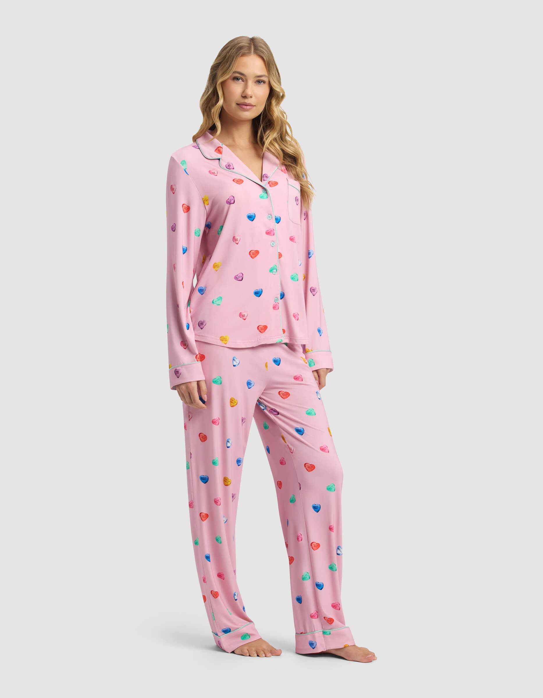 A woman with long blonde hair models the Cozy Earth Women's Bamboo Stretch-Knit Long Sleeve Pajama Set in pink with a colorful heart print, featuring a button-up top and matching pants, against a plain light gray background.|Color:Primrose Sleepy Hearts