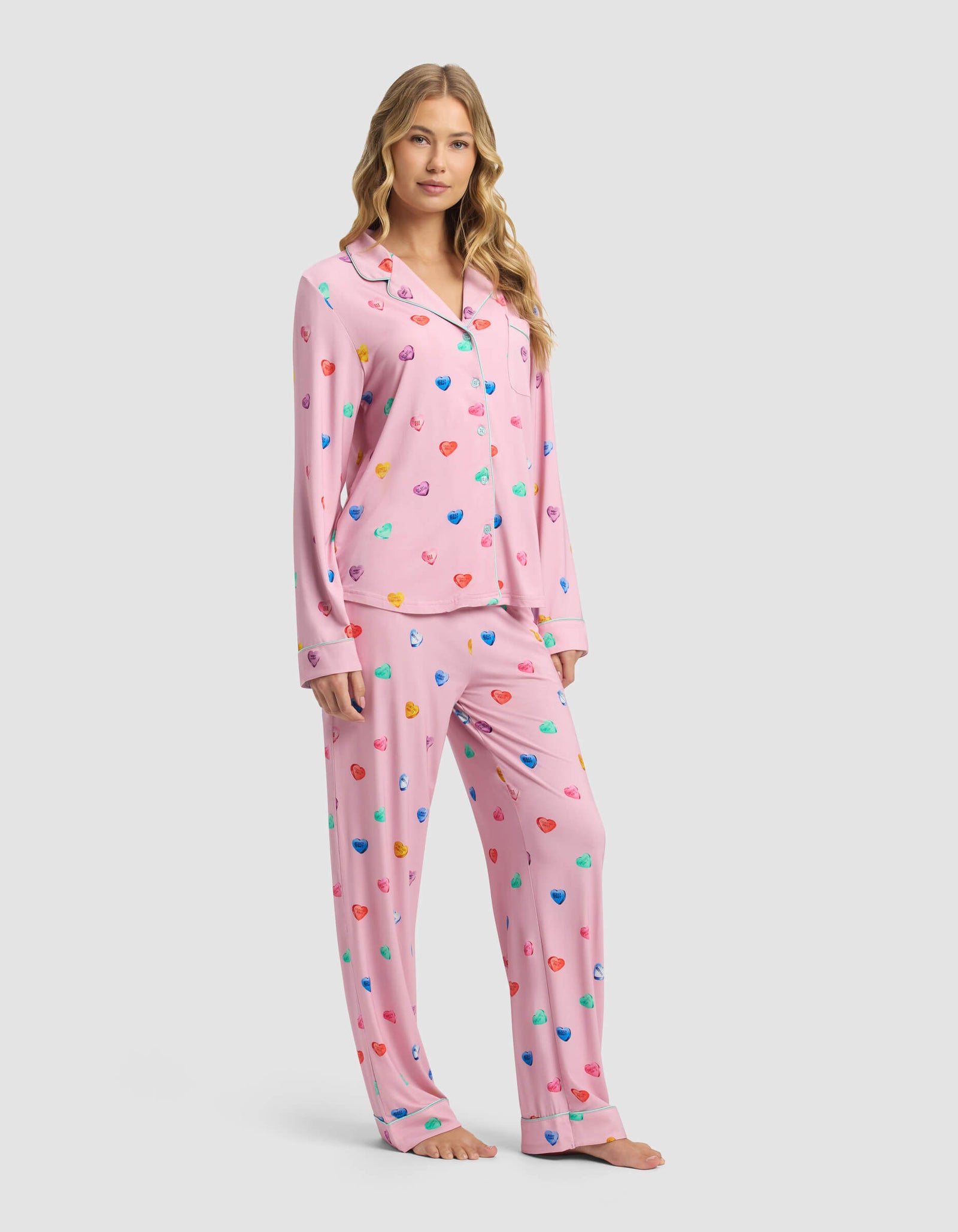A woman with long blonde hair models the Cozy Earth Women's Bamboo Stretch-Knit Long Sleeve Pajama Set in pink with a colorful heart print, featuring a button-up top and matching pants, against a plain light gray background.
