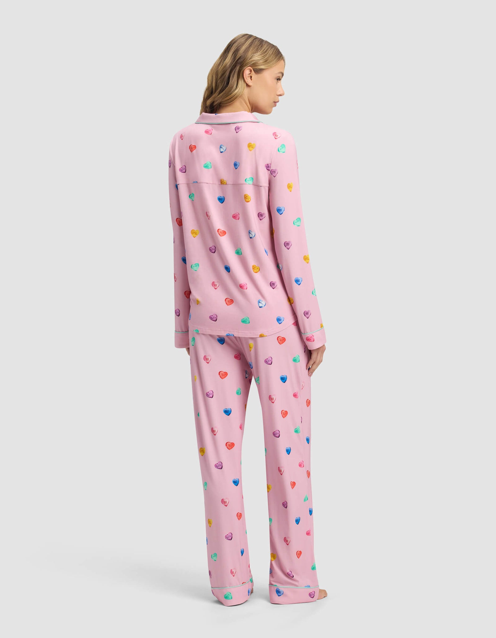 A woman with blonde hair wears the Cozy Earth Women's Bamboo Stretch-Knit Long Sleeve Pajama Set in pink with colorful heart patterns, standing with her back to a plain light gray background.