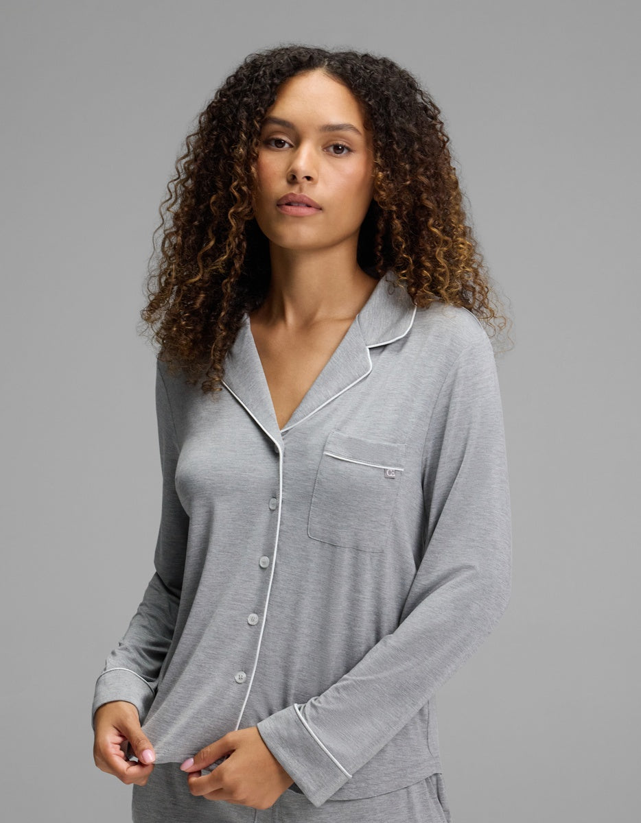 A woman with curly hair stands against a plain gray background, looking at the camera with a neutral expression while wearing the Cozy Earth Women’s Bamboo Stretch-Knit Long Sleeve Pajama Set - Petite in light gray, featuring white piping and buttons.