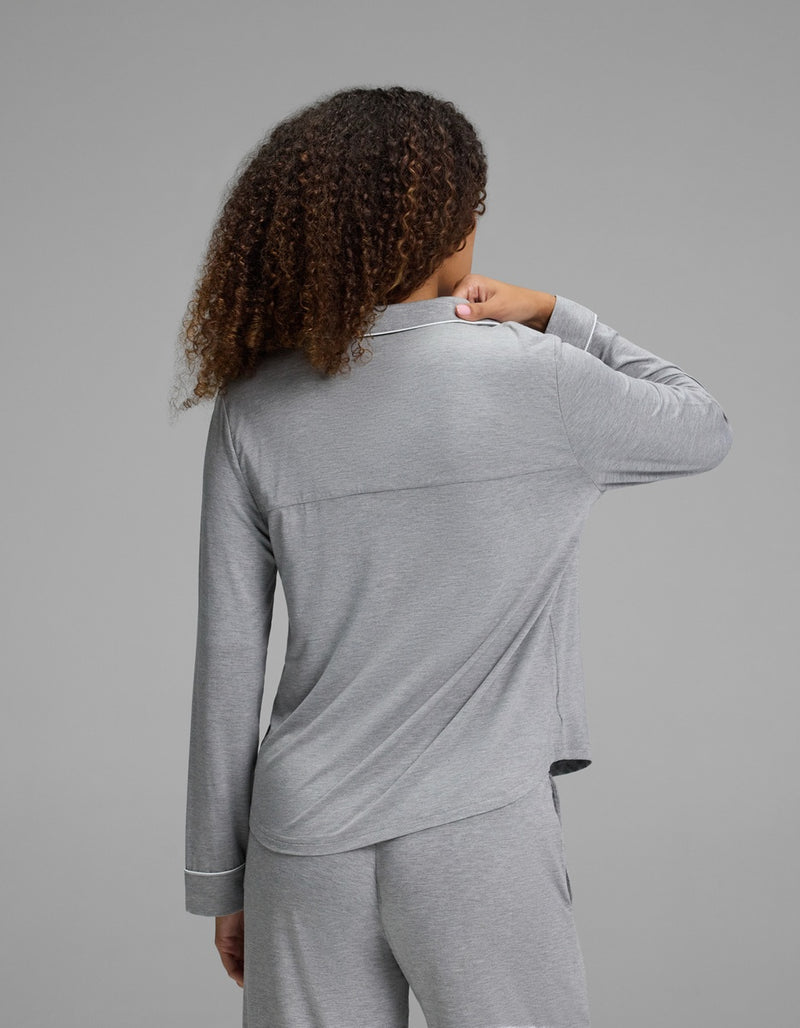 A person with curly hair, seen from behind, wears the Cozy Earth Women’s Bamboo Stretch-Knit Long Sleeve Pajama Top - Petite in light gray, paired with matching pants, against a plain gray background.