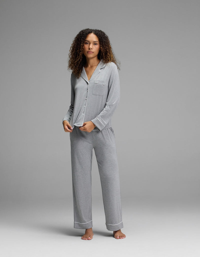 A person with curly hair stands barefoot on a gray background, wearing the Cozy Earth Women’s Bamboo Stretch-Knit Long Sleeve Pajama Top - Petite in light gray, paired with matching long pants.