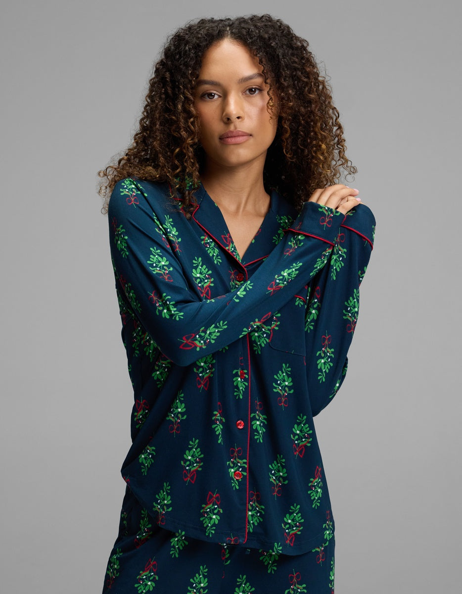 A woman wearing a Cozy Earth Women’s Bamboo Stretch-Knit Long Sleeve Pajama Top in dark blue with a mistletoe and red ribbon print and red piping.