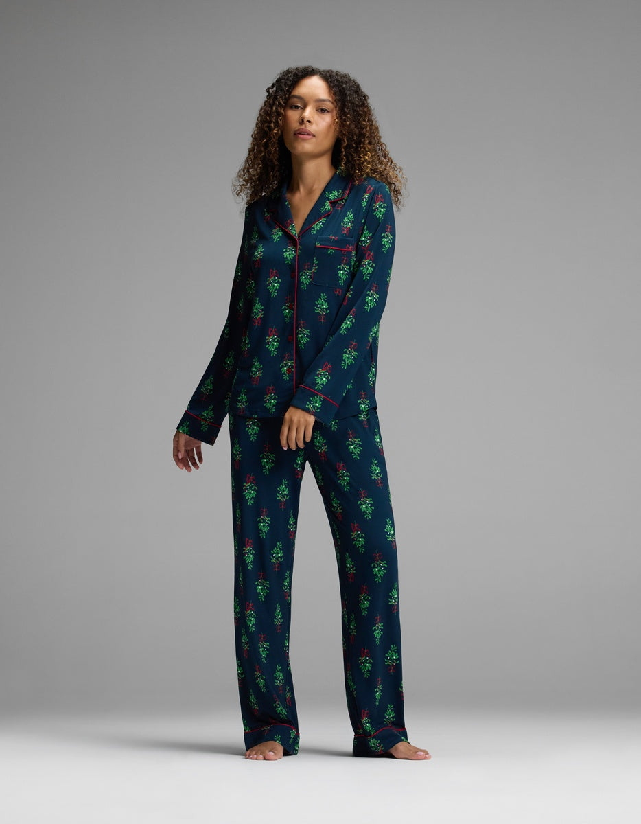 A person wearing a Cozy Earth Women’s Bamboo Stretch-Knit Long Sleeve Pajama Top and matching trousers in a dark blue fabric with a pattern of green Christmas trees and red bows.