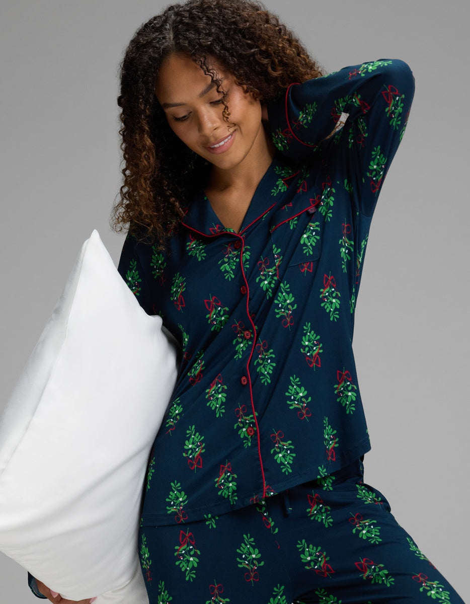 A woman wears a Cozy Earth Women’s Bamboo Stretch-Knit Long Sleeve Pajama Top in navy blue with a mistletoe and red ribbon print and red piping, holding a white pillow.