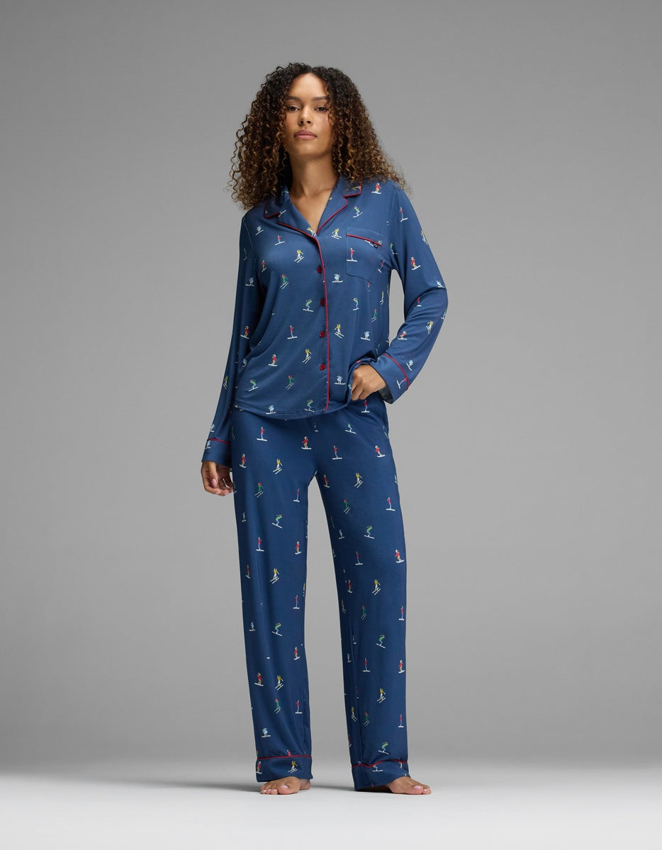 A woman wearing a navy blue Cozy Earth Women’s Bamboo Stretch-Knit Long Sleeve Pajama Top and matching pants, featuring a pattern of small figures and red piping detail on the long-sleeved button-up top.