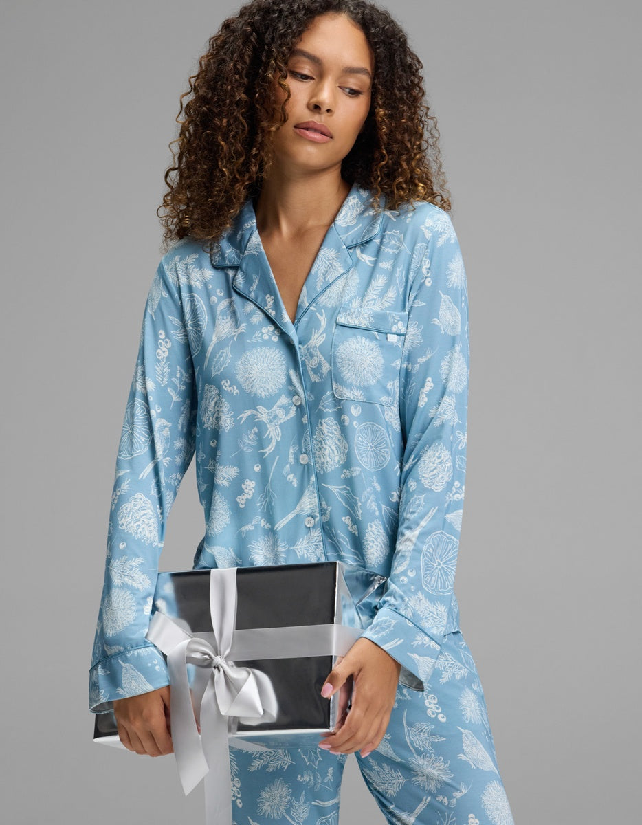 A person wearing a light blue, white botanical print Cozy Earth Women’s Bamboo Stretch-Knit Long Sleeve Pajama Top, holding a black gift box tied with a silver ribbon.