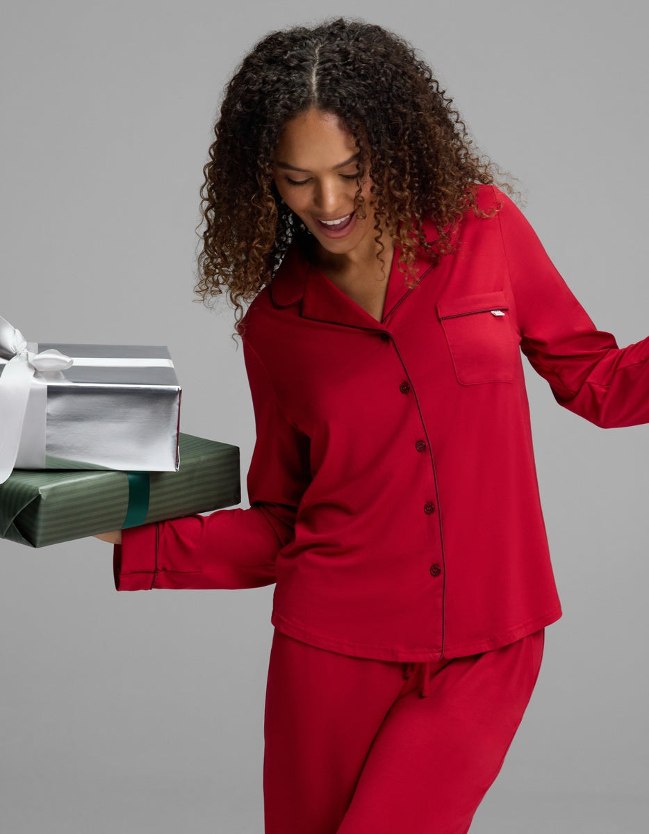 A woman wearing a red Women’s Bamboo Stretch-Knit Long Sleeve Pajama Set with dark piping holds two wrapped gift boxes.