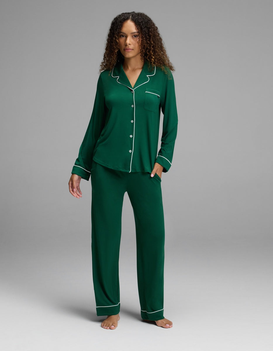 A person wearing a dark green Cozy Earth Women’s Bamboo Stretch-Knit Long Sleeve Pajama Set, consisting of a long-sleeved button-down top with white piping and matching pants.