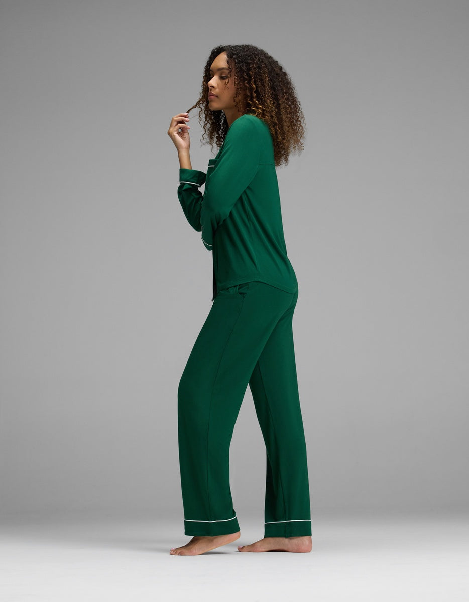 A woman wears a dark green Cozy Earth Women’s Bamboo Stretch-Knit Long Sleeve Pajama Set featuring matching pants with white piping detail.