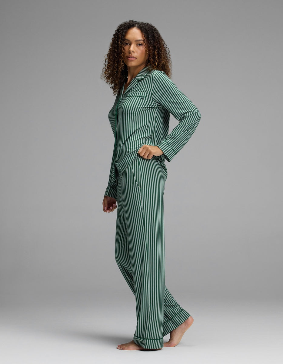 A person wears a Women’s Bamboo Stretch-Knit Long Sleeve Pajama Set, featuring a long-sleeved button-up shirt and matching pajama pants with a dark green and black vertical striped pattern.