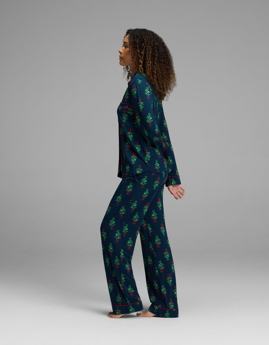 A person wearing a Cozy Earth Women’s Bamboo Stretch-Knit Long Sleeve Pajama Set in a long-sleeved button-up top and matching pants featuring a dark blue pattern of small green Christmas trees and red bows.
