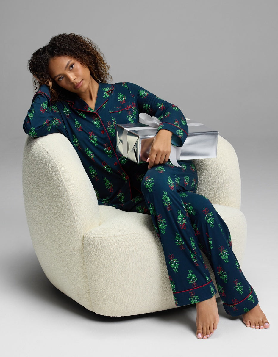 A person wearing a navy blue mistletoe-print Cozy Earth Women’s Bamboo Stretch-Knit Long Sleeve Pajama Set with red piping, sitting in a white armchair and holding a silver gift box.