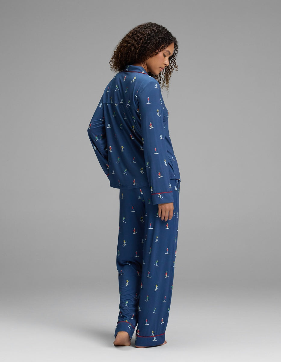 A woman wears a Cozy Earth Women’s Bamboo Stretch-Knit Long Sleeve Pajama Set in navy blue, featuring a long-sleeved button-up top, matching pants, a colorful winter sports print, and red piping.
