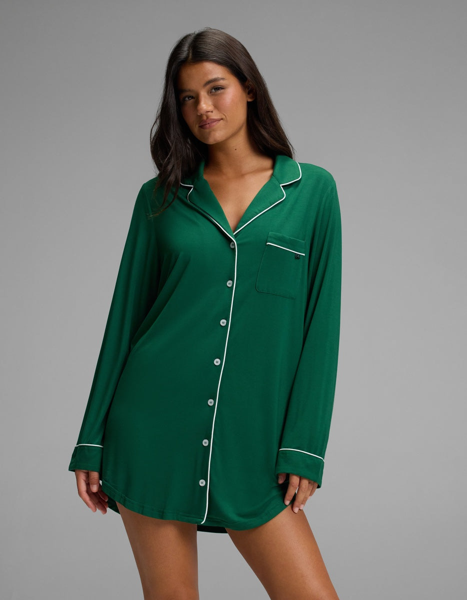 A woman wearing a dark green, long-sleeved Cozy Earth Women’s Bamboo Stretch-Knit Long Sleeve Sleep Dress with white piping trim.