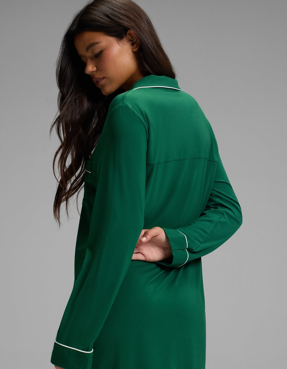 A person wearing a Cozy Earth Women’s Bamboo Stretch-Knit Long Sleeve Sleep Dress in emerald green with white piping trim along the collar and cuffs.